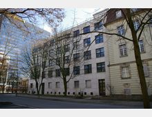 Straßenansicht eines mehrstöckigen Gebäudekomplexes mit beige-weißer Fassade, großen rechteckigen Fenstern und angrenzendem Altbau. Im Hintergrund ist ein modernes Hochhaus mit gläserner Fassade zu sehen, während kahle Bäume im Vordergrund den Blick teilweise verdecken.