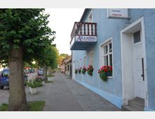 Straßenszene mit einem Gebäude mit blauer Fassade und weißen Fenstern, an denen Blumenkästen mit roten Blumen hängen. Daneben verläuft ein Gehweg mit Kopfsteinpflaster, gesäumt von Bäumen und geparkten Autos.