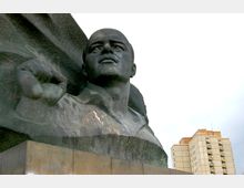 Bronzeskulptur eines Mannes mit erhobenem Arm auf einem Sockel, im Hintergrund ein modernes mehrstöckiges Gebäude vor bedecktem Himmel.