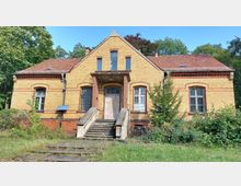 Zweistöckiges, gelbes Backsteingebäude mit rotem Satteldach und dekorativen roten Ziegelakzenten. Das Gebäude hat einen zentralen Eingang mit Steintreppe und ist von Gras und Sträuchern umgeben, im Hintergrund sind Bäume zu sehen.