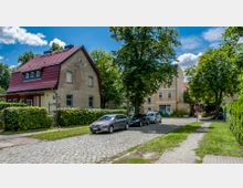 Wohnstraße mit Kopfsteinpflaster in einem Vorstadtviertel, umgeben von Wohnhäusern, darunter ein Haus mit rotem Satteldach und ein beige gestrichenes Gebäude im Hintergrund. Am Straßenrand parken mehrere Autos, und die Szene ist von Bäumen und Grünflächen umrahmt.