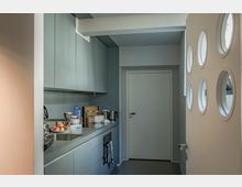 Kleine moderne Küche mit grauen Schränken, einer Arbeitsfläche mit Spüle und Haushaltsgeräten wie Wasserkocher und Ofen. Im Vordergrund ist eine weiße Tür mit kreisförmigen Fenstern sichtbar.