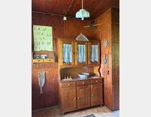 Holzvertäfelter Innenraum mit einem traditionellen Holz-Buffetschrank mit Glasfront und Spitzenvorhängen, darüber eine Hängelampe und ein kleiner Deckenventilator. An der Wand hängen ein Poster und ein kleines Regal, daneben befinden sich Haken und Küchenutensilien neben einer Tür.
