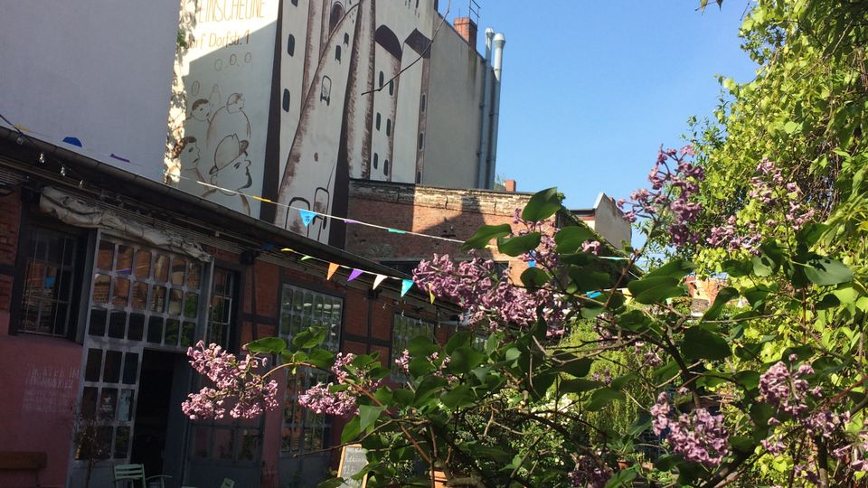 Innenhof zwischen mehrstöckigen Gebäuden mit großen Wandgemälden auf den Fassaden und einer verglasten Werkstattfront. Im Vordergrund stehen blühende Sträucher und weitere Begrünung; über dem Hof hängen bunte Wimpelketten vor blauem Himmel.