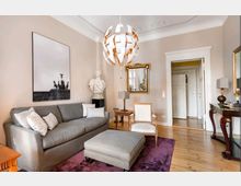 Wohnzimmer in klassischem Stil mit einer grauen Couch, passendem Hocker und einem violetten Teppich. An der Wand hängt ein gerahmtes Kunstwerk, daneben steht eine Büste auf einem Sockel, während ein großer, verzierter Spiegel und eine moderne hängende Deckenleuchte den Raum dekorieren.