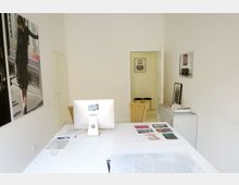 Ein minimalistisches Büro mit einem weißen Schreibtisch, auf dem ein Computerbildschirm, Postkarten und Papier liegen. An der Wand hängen Fotos, und im Hintergrund führt eine offenstehende Tür in einen weiteren Raum mit schlichten Holzhockern und gerahmten Bildern.