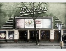 Fassade eines historischen Kinos mit dem Schriftzug „Delphi“ in geschwungener Schrift auf grünem Hintergrund. Darunter befinden sich Filmplakate und mehrere Eingangstüren, mit einem Baum und einem Verkehrsschild im Vordergrund.