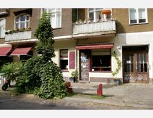 Fassade eines Wohngebäudes mit einer Espressobar im Erdgeschoss, kleinen Balkons mit Pflanzen und einem überstehenden Sonnenschirm. Daneben wächst eine dichte Grünpflanze, und vor der Bar befinden sich beschmierte Poller sowie ein Eingang mit Holztür.