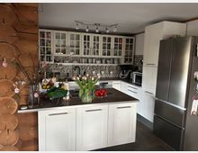 Moderne Küche mit weißen Schränken, einer zentralen Arbeitsfläche und einem großen Edelstahlkühlschrank auf der rechten Seite. Die Rückwand ist mit ornamentierten Fliesen gestaltet, dekoriert mit Frühlingszweigen, Tulpen und Küchenutensilien.