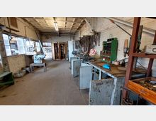 Werkstatt mit alten Arbeitsflächen, Werkzeugen und Maschinen an einer weißen Ziegelwand. Im Hintergrund stehen offene Schränke und Regale, während an der linken Seite große Fenster Licht in den Raum lassen.