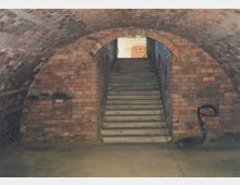 Backstein-Gewölbekeller mit gemauerten Rundbögen und Treppe, die zu einem höher gelegenen Bereich mit Tageslicht führt. Sichtbare Kabel und Leitungen liegen an den Wänden und dem Boden.