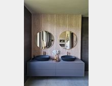 Modernes Badezimmer mit wandhängendem Waschtisch in grauer Farbe, zwei schwarzen Aufsatzwaschbecken und goldenen Wasserhähnen. Die Rückwand ist mit vertikalen Holzpaneelen verkleidet, die zwei runde Spiegel mit goldenen Rahmen hervorheben, während eine dekorative Vase in der Mitte platziert ist.