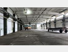 Große, leere Industriehalle mit Betonboden, weißen Wänden und einer Decke mit sichtbaren Stahlträgern. Auf dem Boden stehen zwei Anhänger; Licht fällt durch Fensterreihen an den Seitenwänden ein.