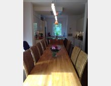 Langer Essbereich in einer modernen Küche mit einem Holztisch, umgeben von geflochtenen Stühlen. Im Hintergrund sind weiße Küchenschränke, eine großzügige Fensterfront mit Blick ins Grüne und eine Hängelampe mit mehreren Leuchtelementen zu sehen; in der Tischmitte steht eine Vase mit einem Blumenstrauß.