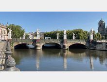 Steinerne Brücke mit dekorativen Skulpturen an den Pfeilern, die über einen Fluss mit ruhigem Wasser führt; im Hintergrund sind Bäume, historische Gebäude und eine Kirche mit Kuppel zu sehen.