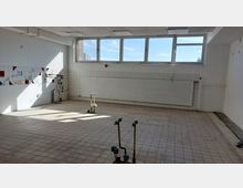 Leerer Raum mit gefliesten Boden- und Wandflächen; an einer Wand befindet sich ein Heizkörper mit geschlossenen Jalousien, darüber mehrere Fenster, die Tageslicht hereinlassen. An der linken Wand ist farbige Wandfliesen-Dekoration sichtbar, und im Raum stehen freiliegende Wasseranschlüsse oder Rohrleitungen.