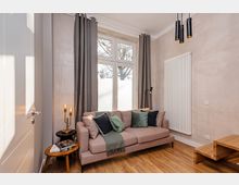 Modernes Wohnzimmer mit hellen Wänden, Holzboden und einem Sofa vor hohen Fenstern mit grauen Vorhängen. An der Wand hängt ein großer weißer Heizkörper; daneben stehen ein kleiner Beistelltisch mit Lampe und ein niedriger Couchtisch, darüber schwarze Pendelleuchten.