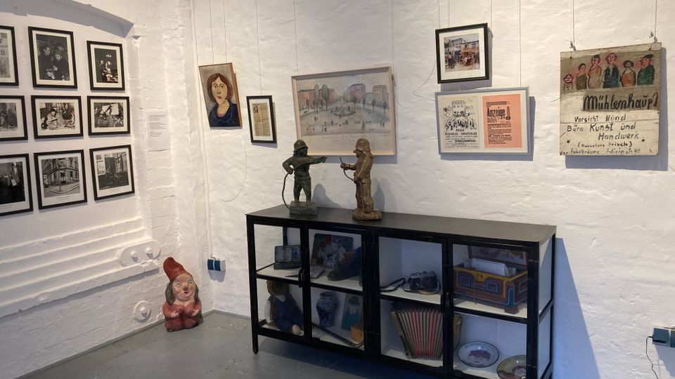 Ausstellungsraum mit weiß verputzten Wänden, an denen gerahmte Fotografien, Zeichnungen und historische Plakate hängen. Davor steht eine schwarze Glasvitrine mit kleinen Objekten und Figuren, auf dem Boden daneben eine Zwergenfigur.