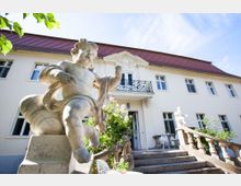 Historisches zweistöckiges Gebäude mit heller Fassade und rotem Dach, einer verzierten Balkontür im Obergeschoss und breiter Treppe mit dekorativen Statuen. Im Vordergrund steht eine marmorene Figur eines Engels, umgeben von Blumen und einem Sitzbereich.