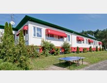 Ein einstöckiges Gebäude mit weißer Fassade, grünem Dach und roten Fenster-Markisen, umgeben von kleinen Büschen und Bäumen. Im Vordergrund steht eine Tischtennisplatte auf einer Wiese, im Hintergrund erstreckt sich ein Wald.