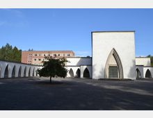Moderne Kirchenanlage mit weißem Putz und gotischen Bögen, umgeben von Arkaden mit Spitzbögen. Im Vordergrund steht ein einzelner Baum auf einem gepflasterten Platz, im Hintergrund ein mehrstöckiges rotes Backsteingebäude.