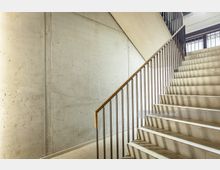 Minimalistisches Treppenhaus mit Betonwänden, einer Treppe aus hellem Stein und einem Geländer aus Metallstäben mit hölzernem Handlauf. Natürliches Licht fällt durch ein Fenster im Hintergrund ein und erzeugt Schattenmuster an der Wand.