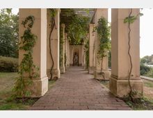 Ein Säulengang mit hellem Mauerwerk, überwachsen von Kletterpflanzen, führt entlang eines gepflasterten Wegs zu einer Statue am Ende des Gangs. Im Hintergrund sind Büsche und Bäume in einer Parklandschaft sichtbar.