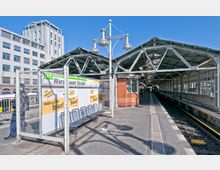 Bahnhof Warschauer Straße in Berlin, mit überdachten Bahnsteigen, einem roten Backsteingebäude und Sitzbänken im Vordergrund; im Hintergrund moderne Architektur sowie Schienen und ein Glasdach.
