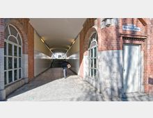 Ein überdachter Fußgängerdurchgang mit gelb gestrichenen Wänden und weißen Kacheln, flankiert von roten Backsteinmauern mit großen Bogenfenstern. Am Ende des Tunnels führen Treppen nach oben, und rechts ist ein Schild mit der Aufschrift „Max-Koch-Passage“ zu sehen.