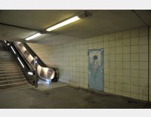 U-Bahn-Station mit Treppe und Rolltreppe auf der linken Seite, gefliesten Wänden in Beige, einer Deckenleuchte und einer abgenutzten blauen Tür auf der rechten Seite.