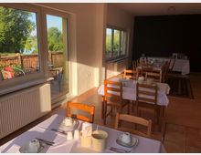 Heller Speiseraum mit Holzmöbeln und weiß gedeckten Tischen, einer großen Fensterfront mit Blick auf eine Terrasse und grüne Landschaft im Hintergrund. Im Raum steht ein Buffet mit Geschirr und Getränken auf einem separaten Tisch.