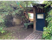 Garten mit einem kleinen gemauerten Schuppen, der eine blaue Holztür und ein Milchglasfenster hat. Links davon befinden sich Heuballen, ein Fahrrad und überwucherte Sträucher, während ein Kopfsteinpflasterweg durch die Szene führt.