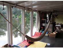Innenraum eines Hausbootes mit großen Fenstern, die auf einen Fluss und Bäume im Hintergrund blicken. Im Vordergrund hängt eine Hängematte neben bunten Kissen und Teppichen, während Regale und Körbe für Stauraum genutzt werden.