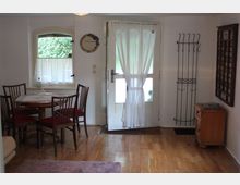 Kleiner Wohnraum mit Holzboden und weiß gestrichenen Wänden. Im Raum befinden sich ein runder Holztisch mit vier Stühlen, ein Fenster mit Vorhang, eine weiße Tür mit durchscheinendem Vorhang und eine Wandgarderobe aus Metall.