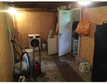 Ein einfacher Kellerraum mit Holzverkleidung an den Wänden und einer sichtbaren Holzbalkendecke. Im Raum befinden sich eine Waschmaschine, ein Staubsauger, Kartons, Plastikflaschen in einem Getränkekasten, sowie weitere Gegenstände wie eine offene Tür, die zu einem angrenzenden Raum führt.