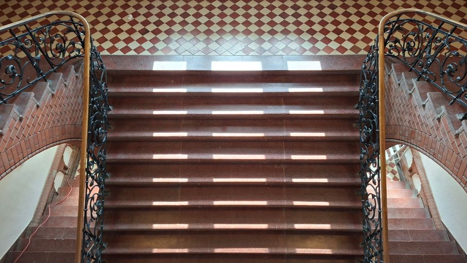 Blick von oben auf ein symmetrisches Innen-Treppenhaus mit breiten braunen Steinstufen und kunstvoll verzierten schwarzen schmiedeeisernen Geländern. Am oberen Podest liegt ein rot-beigefarbener Schachbrettfliesenboden, die Wände sind mit Ziegelbögen eingefasst.