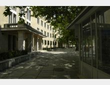 Seitenansicht eines Gebäudeeingangs mit Säulen und großen Fenstern, umgeben von Gehwegen und begrenzten Grünflächen. Rechts im Bild befindet sich ein verglaster Pavillon, während im Hintergrund Bäume Schatten auf die Szenerie werfen.