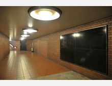 Unterirdischer Gang mit roten Fliesen auf dem Boden und Wänden aus rot-braunem Ziegelstein. Die Decke ist mit runden, hell erleuchteten Lampen ausgestattet, und am Ende des Gangs führt eine Treppe nach oben; eine große schwarze Tafel ist an der rechten Wand befestigt.