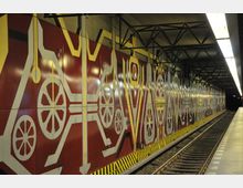 U-Bahn-Station mit künstlerisch gestalteten Wänden in Rot, Weiß und Gelb, die große, radähnliche Muster zeigen. Der Bahnsteig verläuft parallel zu den Gleisen, und die Decke ist mit Metallträgern und Beleuchtungselementen versehen.
