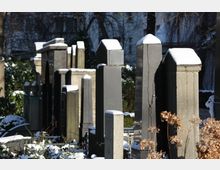 Reihe von Grabsteinen auf einem verschneiten Friedhof, teilweise von winterlichem Sonnenlicht beleuchtet. Im Hintergrund sind Bäume und unscharfe Gebäudefassaden zu erkennen.
