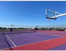 Offenes Basketballfeld mit violettem und rotem Untergrund, umgeben von einem Metallzaun, vor einem Hintergrund mit Wiesen und vereinzelten Gebäuden. Der Himmel ist klar und blau.