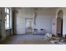 Ein verlassener Raum mit abgeblätterter Farbe an den Wänden, einem kaputten Fußboden und herumliegendem Schutt. Zu sehen sind ein großes Fenster, eine geöffnete Tür, zwei einfache Holzstühle und ein heller Kachelofen an der rechten Wand.