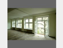 Ein leerer Raum mit hellem Holzfußboden, großen Fenstern auf einer Wandseite und einem geöffneten Fenster mit Blick auf Bäume. Die Wände sind weiß gestrichen, und Heizkörper befinden sich unter den Fenstern.