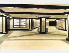 Unterirdischer U-Bahn-Bereich mit einer offenen, rechteckigen Architektur. Weiße Wandfliesen mit schwarzen Akzenten sowie geometrische Säulen und Leuchtflächen prägen das Design; im Hintergrund befinden sich ein Treppenabgang und Werbeplakate.