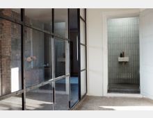 Ein heller Innenraum mit einer modernen Glaswand auf der linken Seite, die einen Bereich mit einer Backsteinwand und einer Kommode mit Blumen in einer Vase abtrennt. Rechts befindet sich eine offene Tür, die in ein minimalistisches Badezimmer mit grünen Wandfliesen, einem kleinen weißen Waschbecken und einer goldfarbenen Armatur führt.