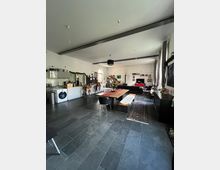 Großer, offener Wohnraum mit modernem Industriedesign, ausgestattet mit einer offenen Küche auf der linken Seite, einem langen Esstisch in der Mitte und einer Sitzecke im Hintergrund. Der Raum hat einen dunklen Steinboden, hohe Decken mit sichtbarem Träger, große Fenster und eine schlicht dekorierte, helle Wandgestaltung.