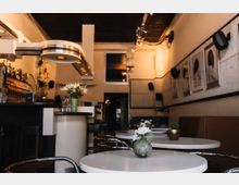 Ein gemütliches Café mit modernen, runden Tischen und eleganten Metallstühlen. An der linken Seite befindet sich eine Bar mit Flaschenregalen, während die Wand rechts dekorative Kunstwerke und Lautsprecher zeigt.