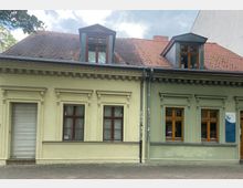 Fassade eines einstöckigen Gebäudes mit gelber und grüner Wand, roten Fenstern mit weißen Rollläden sowie einem Satteldach aus roten Ziegeln und Dachgauben. Der Gehweg davor ist gepflastert, und ein Schild hängt rechts an der grünen Wand.