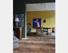 Wohnraum mit klassischem Fischgrätparkett, moderner Einrichtung und farbigen Wänden; im Vordergrund ein grünes Ecksofa neben einer Holzkommode, im Hintergrund eine gelbe Wand mit Gemälde und ein Esstisch mit bunten Stühlen. Deckenstuck und eine minimalistische Hängelampe ergänzen das Ambiente.