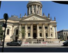 Historisches Gebäude mit neoklassizistischer Fassade, markanten Säulen und detaillierten Skulpturen vor einem blauen Himmel. Vor dem Eingang stehen Sonnenschirme und Absperrungen, während Autos auf der Straße davor parken.